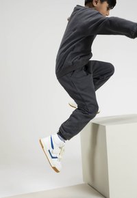 Grijze sweatshirt en bijpassende joggers met ribbelmanchetten, gecombineerd met witte hoge sneakers met blauwe accenten en een rubberen zool, in een sprongetje.