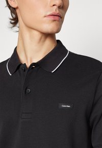 Black polo shirt with a textured fabric, a classic collar, and white accent stitching. Features a Calvin Klein logo patch on the chest.