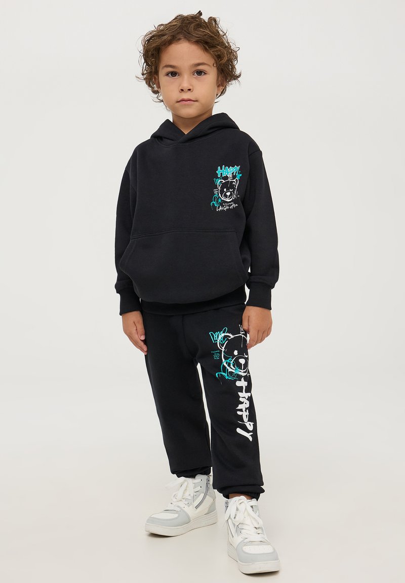 Felpa nera con grafica di un orso turchese stampata sul petto, pantaloni della tuta abbinati con l'orso e il testo "Trippy", abbinati a sneakers bianche.