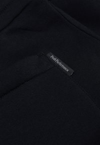 Tessuto nero con una superficie liscia presenta un piccolo etichetta riportante la dicitura "Peak Performance". Il design è semplice e funzionale, senza motivi visibili.