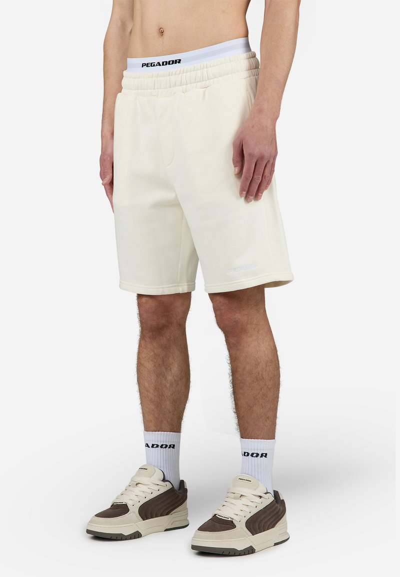 Cremefarvede shorts med elastisk talje, diskret logo detalje, lige ben design og blød tekstur, parret med neutralt farvede sneakers.