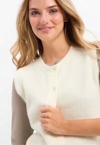 Cremfarbener Strickwesten mit geripptem Muster, runder Ausschnitt und zwei Knopfverschlüssen vorne. Über einem grauen Oberteil getragen.