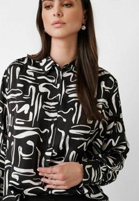 Woman wearing a black button-up shirt with white abstract brushstroke patterns, long dark hair, pearl earrings, and silver rings on her fingers.