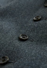 Dunkelgrauer Stoff mit einer sanften Textur, der runde, dunkle Knöpfe mit einem Blumenmuster aufweist. Nahaufnahme der Knopfdetails auf dem Kleidungsstück.