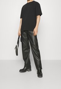 Černé oversized tričko z bavlny, spárované s černými koženými kalhotami s volným střihem a robustními černými botami. Drží malou černou tašku.
