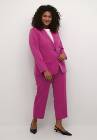 Completo su misura fucsia con blazer a un bottone e pantaloni cropped, abbinato a un top bianco. La modella indossa scarpe col tacco nere.