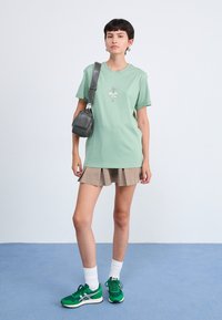 Mintgrön t-shirt med grafiskt tryck på framsidan, ihop med en beige plisserad kjol. Modellen bär gröna sneakers och har en liten grå väska.
