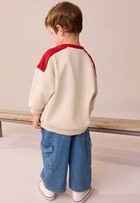 Kind in een crèmekleurige sweatshirt met rode accenten, gecombineerd met brede blauwe denim broeken en witte sneakers. Eenvoudig ontwerp met ribbelmanchetten.