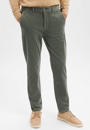 Grüne Cordhosen mit geradem Schnitt, versehen mit einem Knopfverschluss und seitlichen Taschen, kombiniert mit hellbraunen Sneakers.