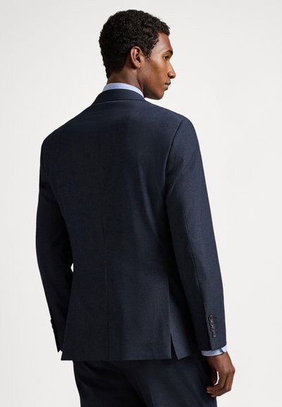 Vīrietis tumši zilos uzvalkā un gaiši zilā krekliņā, redzams no muguras, skatās uz sāniem pret vienmuļu gaišu fonu.