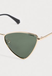 Trokutaste zelene leće u zlatnom metalnom okviru Polaroid sunčane naočale s crnim vrhovima na bijeloj pozadini.