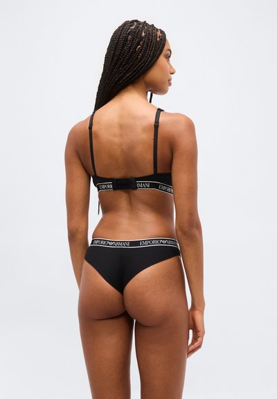 Γυναίκα με πλεξούδες που φοράει μαύρο σουτιέν Emporio Armani και αντίστοιχα εσώρουχα, εμφανιζόμενη από πίσω σε απλό ανοιχτό φόντο.