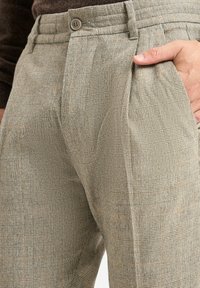 Man met hand in pocket, gekleed in beige geruite broek met knopen en voorplooien, gecombineerd met een donkerbruine trui.