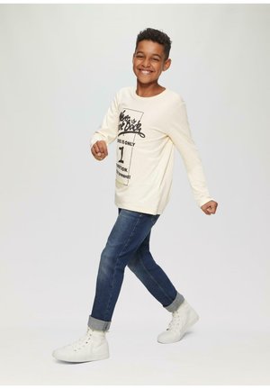 Crème lange mouwen shirt met zwarte grafische print, gedragen met blauwe jeans en witte sneakers. De outfit heeft opgerolde denimmanchetten.