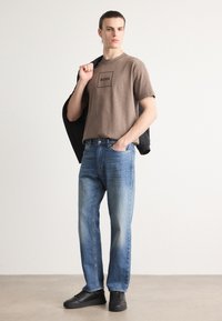 Brun t-skjorte med svart "BOSS" logo, kombinert med lyseblå denimjeans og svarte sneakers. Modellen holder en svart jakke over den ene skulderen.