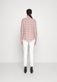 Röd och grå rutmönstrad långärmad skjorta med knäppta manschetter, bärs över vita slim-fit byxor och svarta ankelstövlar. Enfärgad bakgrund.