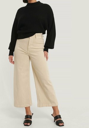 Personne portant un pull en maille noir, un pantalon large beige coupé au-dessus de la cheville et des sandales à talons noires, debout devant un fond uni.