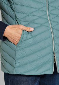 Hand in der Tasche einer hellblauen gesteppten Jacke mit diagonaler Naht und silbernem Reißverschluss, getragen über einem dunklen Ärmel und blauen Jeans.