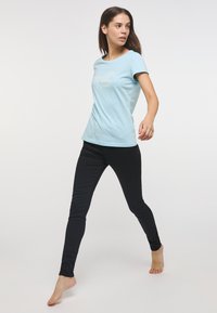 Šviesiai mėlynas trumpomis rankovėmis marškinėliai su apvalia iškirpte ir grafiniu dizainu priekyje, derinami su priglundančiais juodais džinsais. Dėvintis basas modelis judesyje.