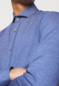 Blauwe overhemd met knoopsluiting, gemaakt van textiel met een subtiel patroon, voorzien van een puntige kraag en twee grijze knopen op de sluiting.