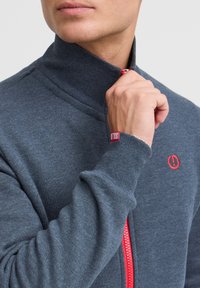 Grau zip-up Sweatshirt mit hohem Kragen, rotem Reißverschluss und rotem Logodetail auf der Brust und dem Ärmel, aus einem weichen, strukturierten Stoff gefertigt.