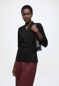 Čierny zavinovací top s dlhými rukávmi, detailom uzla v páse, v kombinácii s bordovými nohavicami a malou čiernou kabelkou.