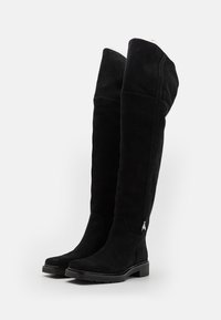 Las botas de ante negras hasta la rodilla presentan una textura suave, un ligero tacón y un sutil acento metálico cerca del tobillo. Diseño elegante y ajustado.