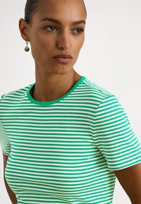 Žena s vlasmi stiahnutými dozadu, ktorá má na ušiach okrúhle zlaté náušnice a nosí zeleno-bielo pruhované tričko s krátkym rukávom, mierne sa pozerá bokom.