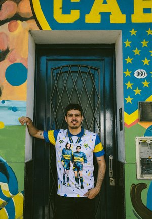 Witte trui met blauwe kraag en gele accenten, met twee voetballers. De achtergrond bevat een blauwe deur met geometrische muurtekeningen.