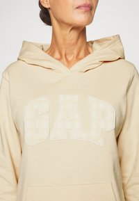 Beige fleece hoodie met een voorvak in de vorm van een kangoeroezak en een geruit "GAP"-logo in lichtgele en witte tinten erop geborduurd. Capuchonmodel.