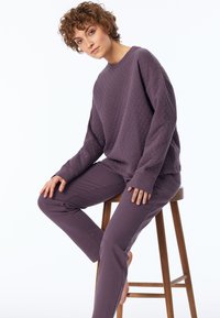 Paarse, gewatteerde sweatshirt met lange mouwen en geribbelde manchetten, gecombineerd met bijpassende slim-fit broeken, allemaal in een zachte stof. Zittend op een houten kruk.