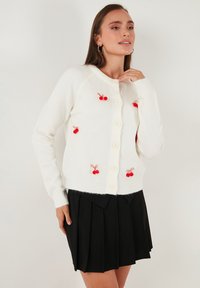Witte gebreide cardigan met rode kersborduring, voorzien van een ronde hals en knoopsluiting. Gecombineerd met een zwarte geplooide rok.