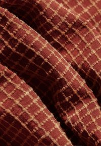 Strukturierter Stoff in Ziegelrot mit einem Karomuster, das helle Quadrate zeigt. Das Material wirkt weich mit einer gewebten Textur.