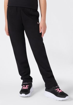 Ben med sorte joggebukser med liten logo og svarte sneakers med rosa snørebånd og hvite såler som står på et hvitt gulv.