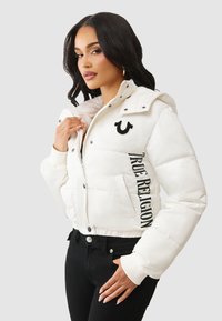 Biała kurtka puchowa o dopasowanym kroju, z kapturem, zamek błyskawiczny, kieszenie boczne oraz czarny napis "TRUE RELIGION" z boku.