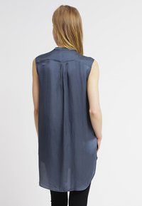 Ärmellose marineblaue Bluse aus einem weichen, strukturierten Stoff. Verfügt über eine Rückenfalte und einen abgerundeten Saum, mit einem dezenten Glanz. Figurbetonte Passform.