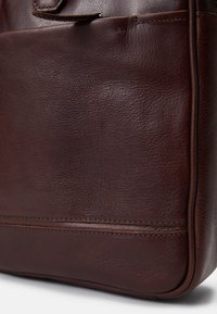 Barna bőrtáska, amely texturált felülettel, látható varrásokkal és karcsú profillal rendelkezik; elöl egy fedélzsebet tartalmaz.