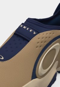 Κοντινό πλάνο ενός μπεζ και ναυτικού αθλητικού παπουτσιού με την επιγραφή "OAKLEY" στο ναυτικό ύφασμα στη μύτη.