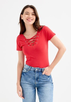 Mujer joven sonriente, vestida con una camiseta roja de manga corta con cordones cruzados en el escote y jeans azules de tiro alto.