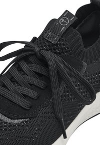 Schwarzer Sportschuh mit strukturiertem Mesh-Obermaterial, runden Schnürsenkeln, gepolstertem Schaftrand und einer weißen Gummisohle mit einem leichten Absatz.