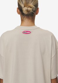 Beiges, übergroßes T-Shirt mit rundem Kragen. Verfügt über ein rosa gesticktes Etikett "Most Alive" am hinteren Halsausschnitt. Glatte Textur.