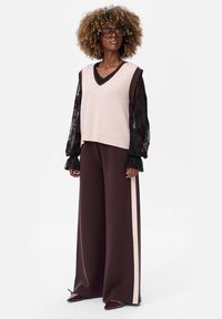 Beżowy dzianinowy kamizelka noszona na czarnej półprzezroczystej bluzce z długimi rękawami, zestawiona z szerokimi brązowymi spodniami z białym paskiem po boku. Błyszczące czerwone czółenka w szpic.