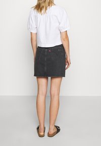 Mini-jupe en denim noir avec deux poches arrière et une étiquette logo rouge. Associée à un haut court blanc à manches courtes et des sandales foncées.