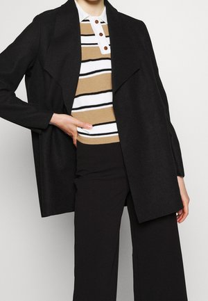 Persoon draagt een zwarte open jas, beige wit en zwart gestreept poloshirt met knopen en zwarte wijde broekspijpen, hand rust op de heup.