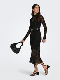 Vestido de encaje negro con mangas largas ajustadas y cuello alto, con un diseño transparente. Combinado con un cinturón negro y un bolso negro. Tacones negros con tiras.