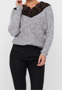 Pull gris tricoté avec un col en V profond orné de dentelle noire au niveau du col. Manches longues et texture côtelée. Porté avec un pantalon noir.