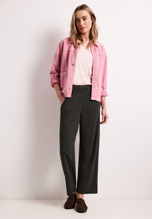 Frau steht an einer weißen Wand, trägt eine hellrosa Jacke, weißes Hemd, schwarze weit geschnittene Hose und braune Loafer, mit einer Hand in der Tasche.