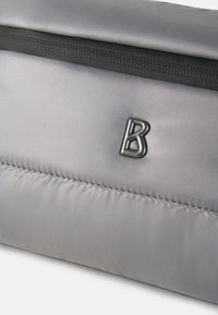 Sivá polstrovaná taška s textúrovaným povrchom, čiernym zipsom a razeným logom 'B'. Hladký materiál s viditeľnými ševmi.