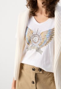 Bílý bavlněný tričko s dekorativním designem křídel ozdobených flitry v modré, zlaté a stříbrné barvě; kombinováno se světle hnědou sukní s vysokým pasem.