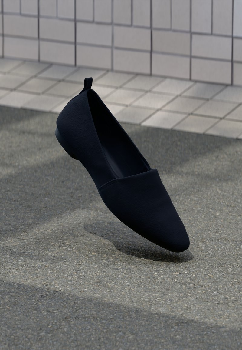 Chaussure plate noire à bout pointu flottant au-dessus d’un bitume gris près d’un mur carrelé avec une petite languette au talon.